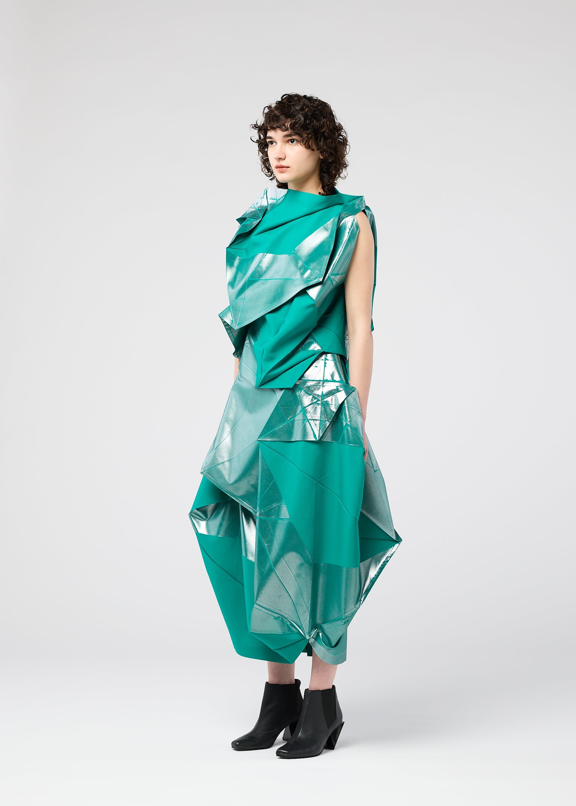 Zoomed view A person with curly hair models the 132 5. ISSEY MIYAKE NO.17 FOIL: a sculptural green geometric outfit with metallic silver accents and folding structure, styled with black ankle boots, standing sideways against a plain white background.