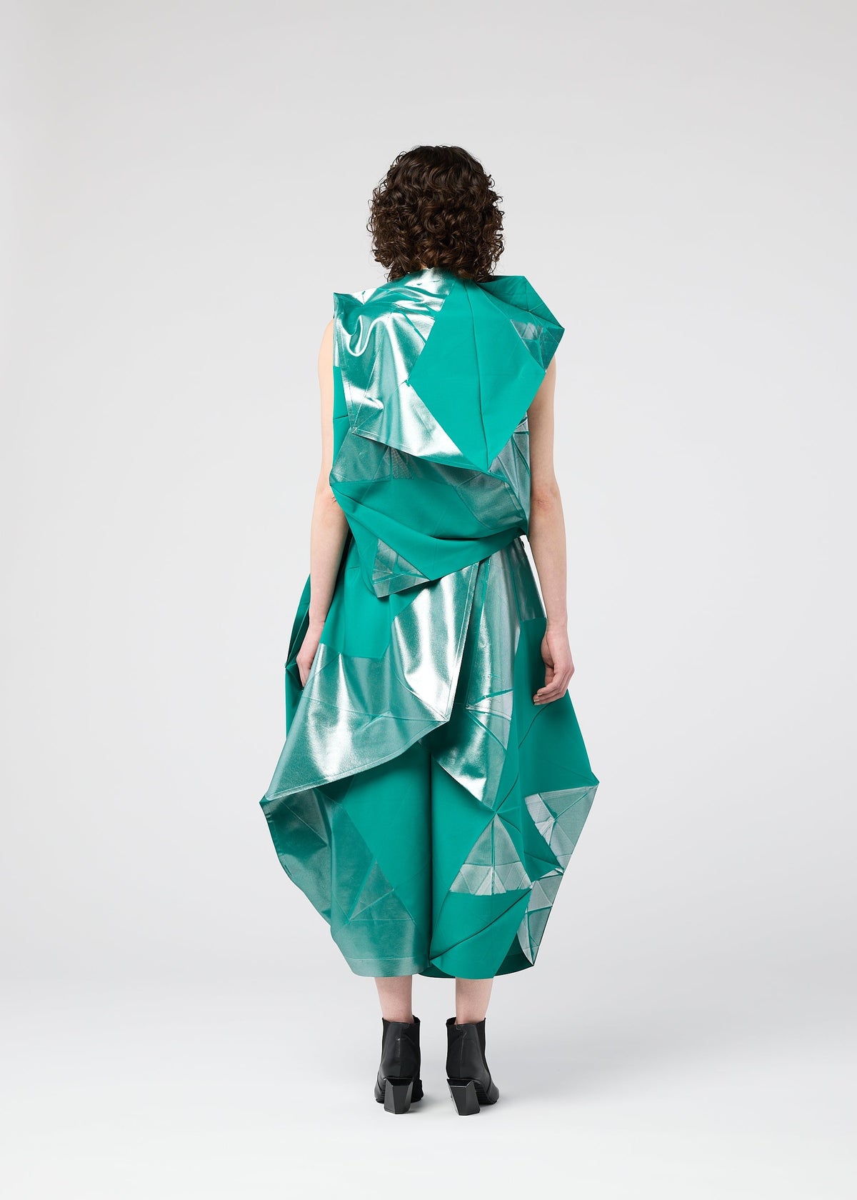 A person with curly hair models the NO.17 FOIL dress by 132 5. ISSEY MIYAKE, a futuristic green and silver geometric design with an innovative folding structure, seen from the back against a plain light background, paired with black ankle boots. view 7 of 10