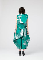 A person with curly hair models the NO.17 FOIL dress by 132 5. ISSEY MIYAKE, a futuristic green and silver geometric design with an innovative folding structure, seen from the back against a plain light background, paired with black ankle boots.