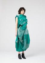 A person with curly brown hair wears the 132 5. ISSEY MIYAKE NO.17 FOIL dress—shiny teal with innovative folding and geometric angles—paired with black ankle boots, standing against a plain light gray background.