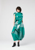 A person with curly hair wears the NO.17 FOIL dress by 132 5. ISSEY MIYAKE—a shiny teal design with innovative angular folds, creating a three-dimensional silhouette—paired with black ankle boots against a plain light gray background.