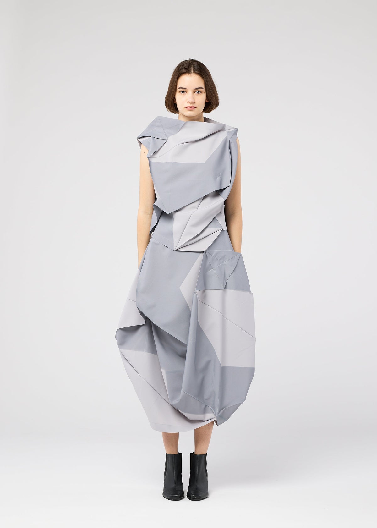 A woman stands against a plain white background wearing the NO.17 PRINT dress by 132 5. ISSEY MIYAKE with black ankle boots, arms relaxed at her sides, looking directly at the camera. view 5 of 9