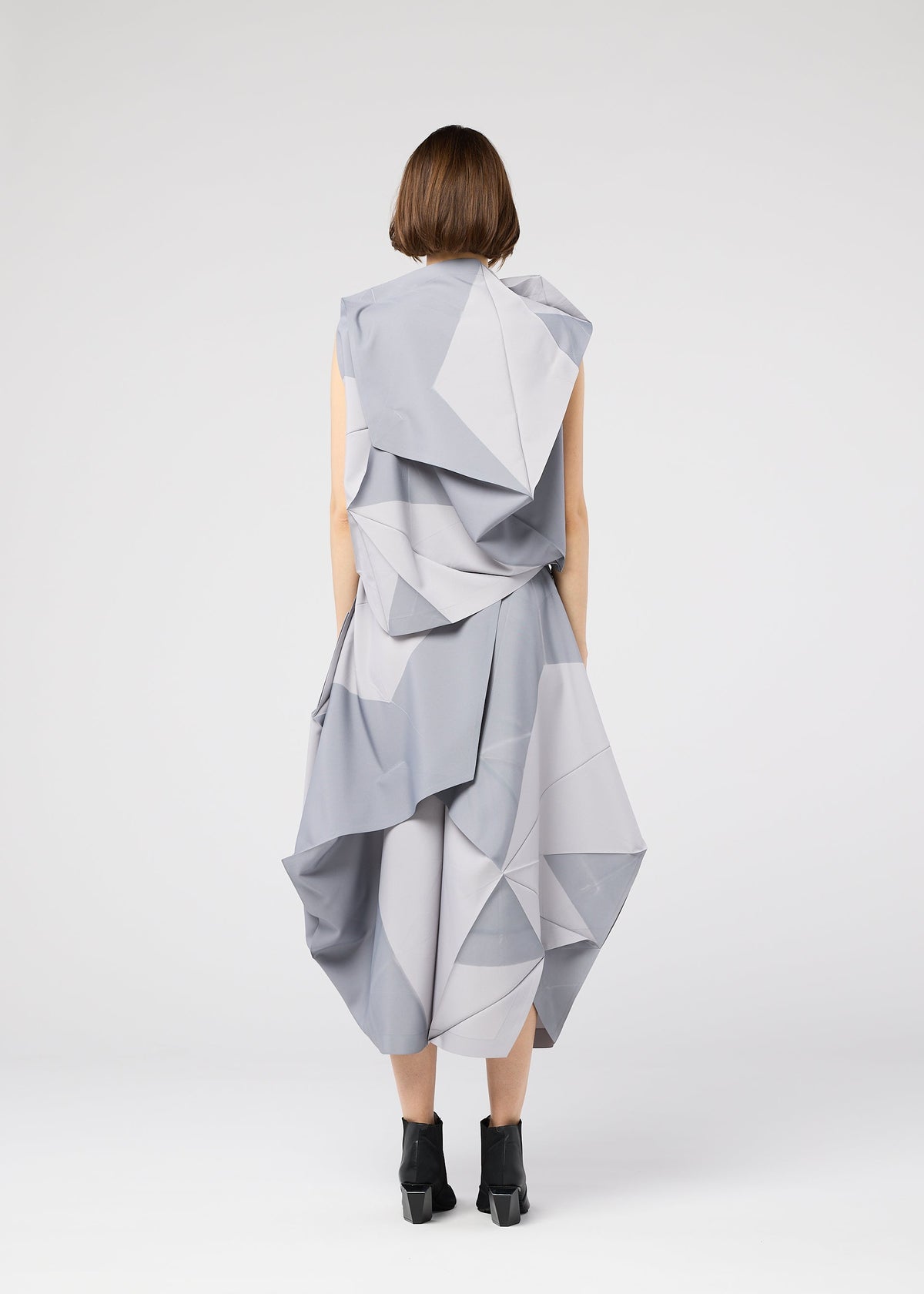 A person with short brown hair, seen from behind, wears the 132 5. ISSEY MIYAKE NO.17 PRINT, a light gray geometric origami-inspired dress with folding structure and black ankle boots against a plain white background. view 7 of 9