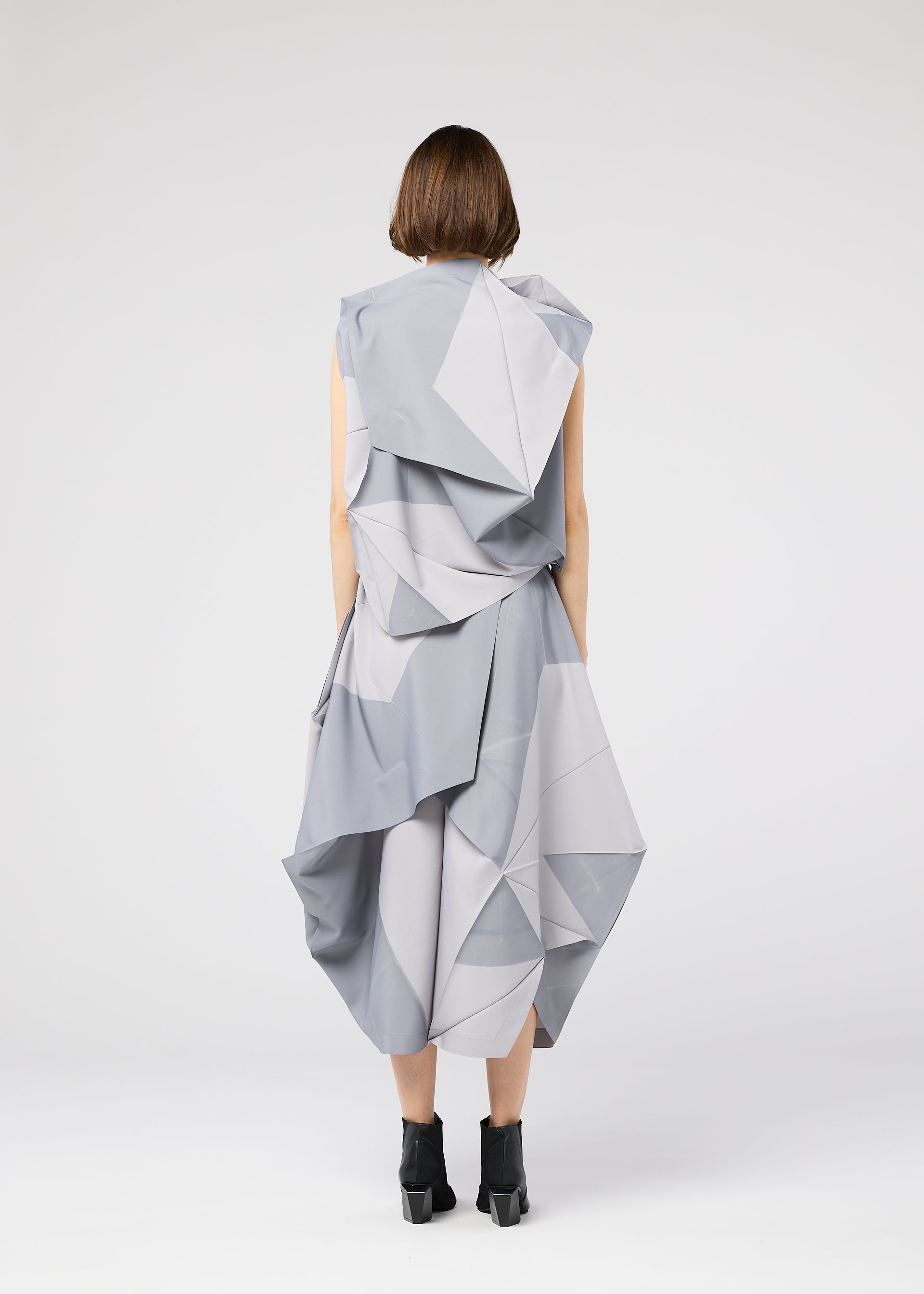 Zoomed view A person with short brown hair, seen from behind, wears the 132 5. ISSEY MIYAKE NO.17 PRINT, a light gray geometric origami-inspired dress with folding structure and black ankle boots against a plain white background.