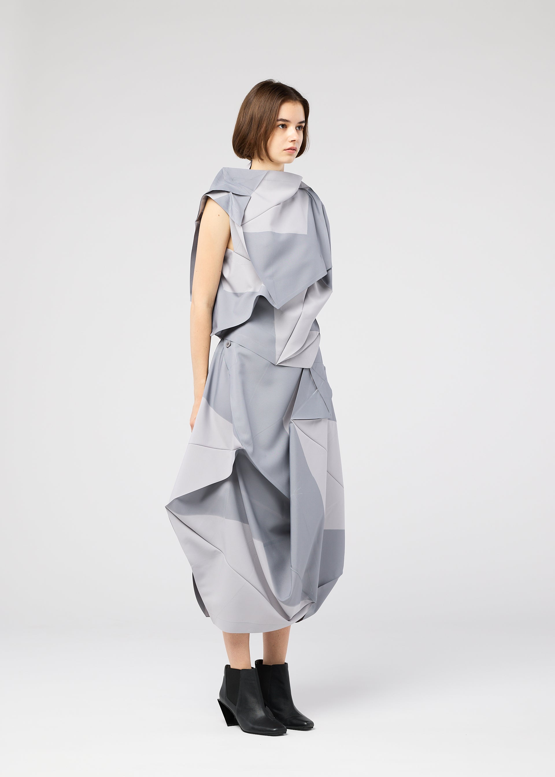 Zoomed view A woman with short brown hair stands in profile, wearing the NO.17 PRINT by 132 5. ISSEY MIYAKE—a light gray, geometric outfit with layered, angular folds—and black ankle boots, set against a plain white background.