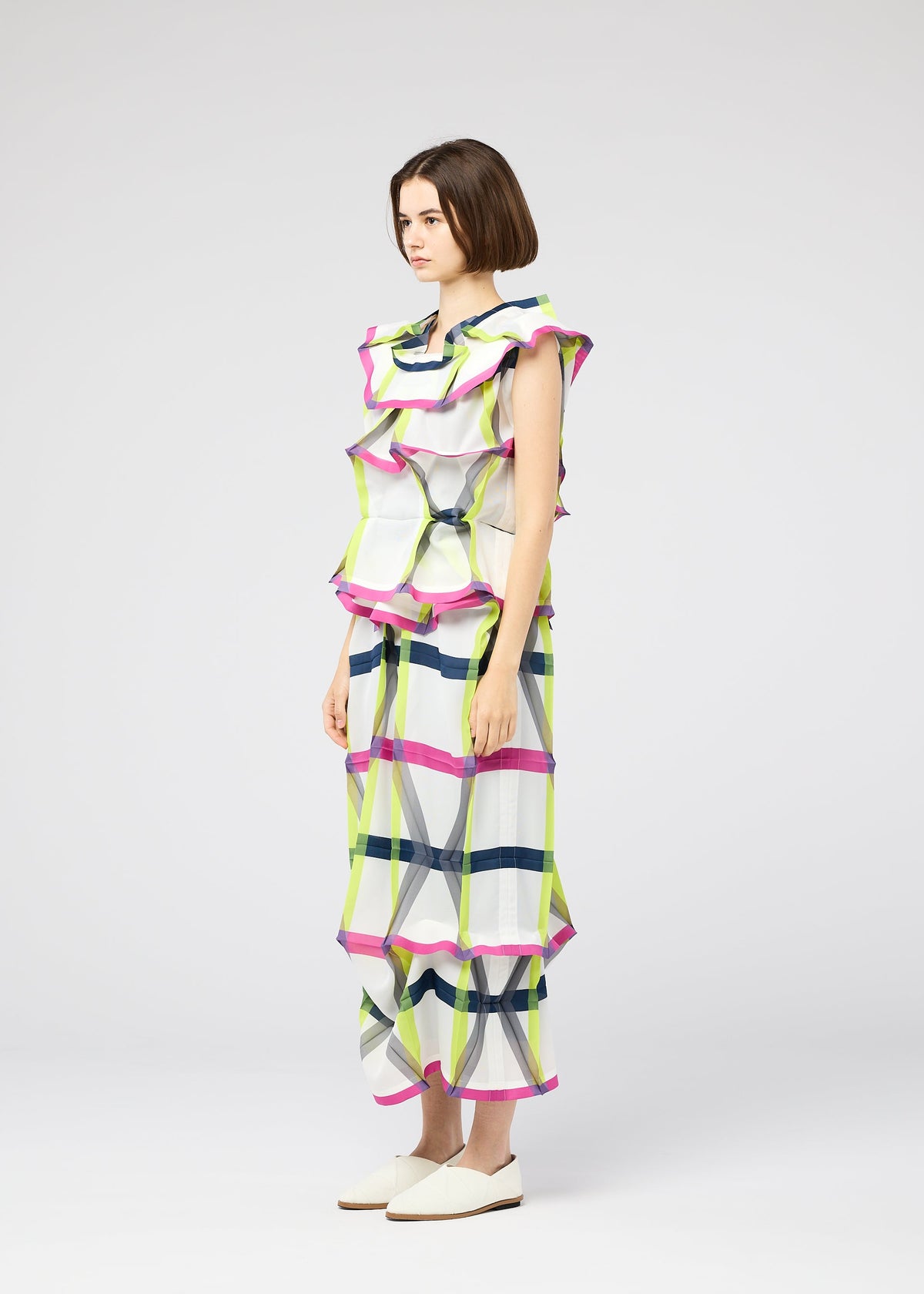 A person stands in profile wearing the 132 5. ISSEY MIYAKE NO.6 TRANSLUCENT set— a sleeveless, ruffled plaid top and matching wide-leg pants in white, pink, yellow, and navy blue—paired with white slip-on shoes against a plain light background. view 8 of 11