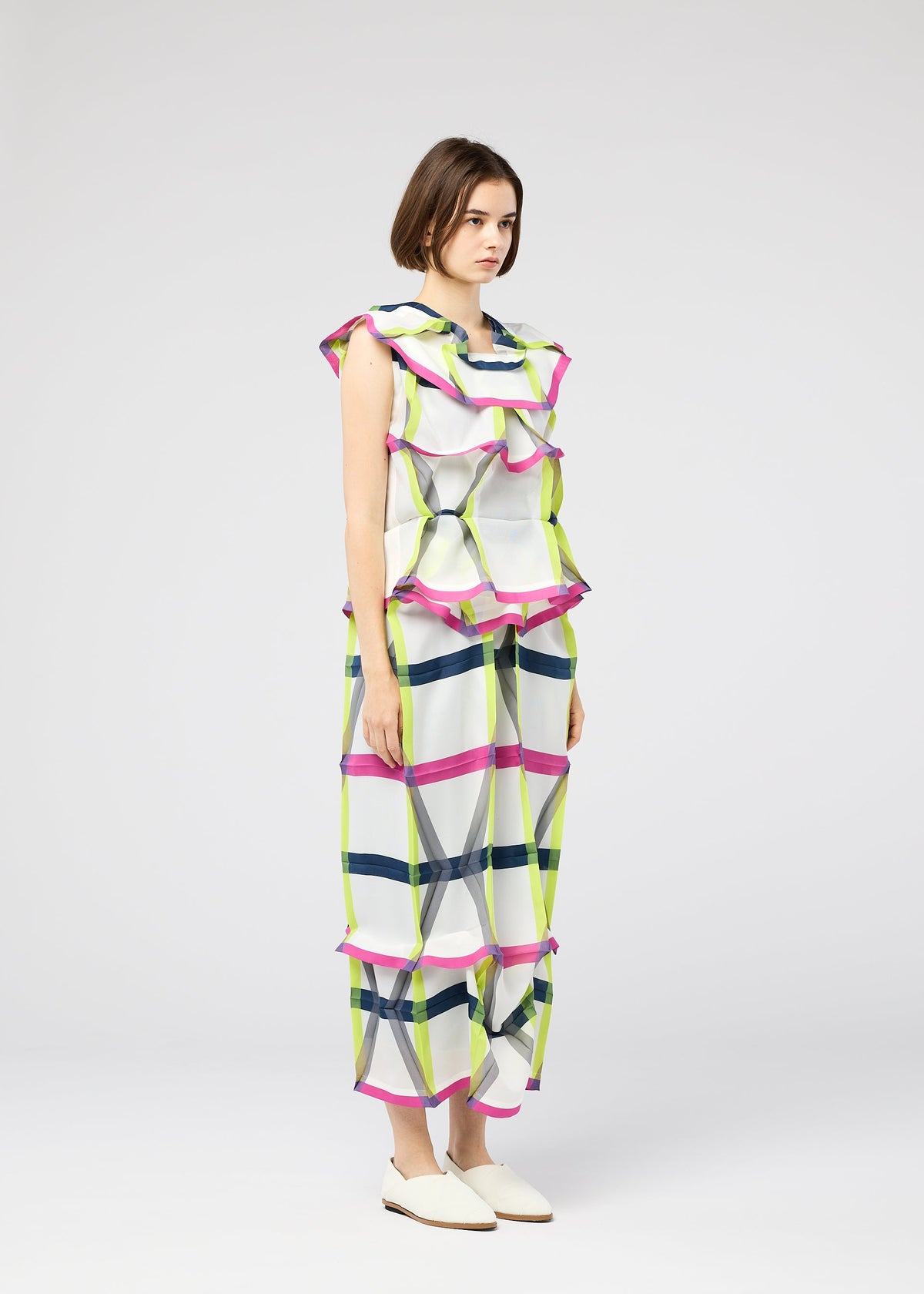 A woman with short brown hair and white flats wears the NO.6 TRANSLUCENT outfit by 132 5. ISSEY MIYAKE—a loose, sleeveless piece with sheer fabric, wide ruffles, and a green, pink, and blue grid pattern on white against a plain gray background. view 10 of 11
