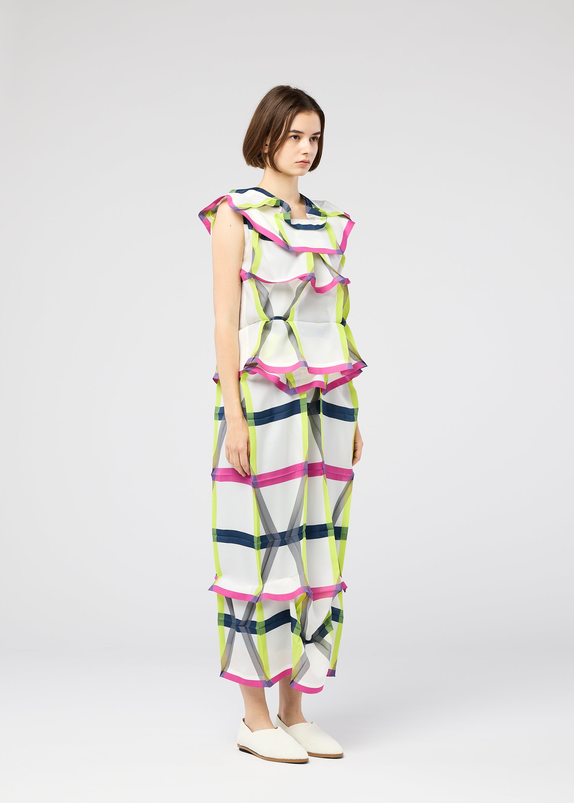 Zoomed view A woman with short brown hair and white flats wears the NO.6 TRANSLUCENT outfit by 132 5. ISSEY MIYAKE—a loose, sleeveless piece with sheer fabric, wide ruffles, and a green, pink, and blue grid pattern on white against a plain gray background.