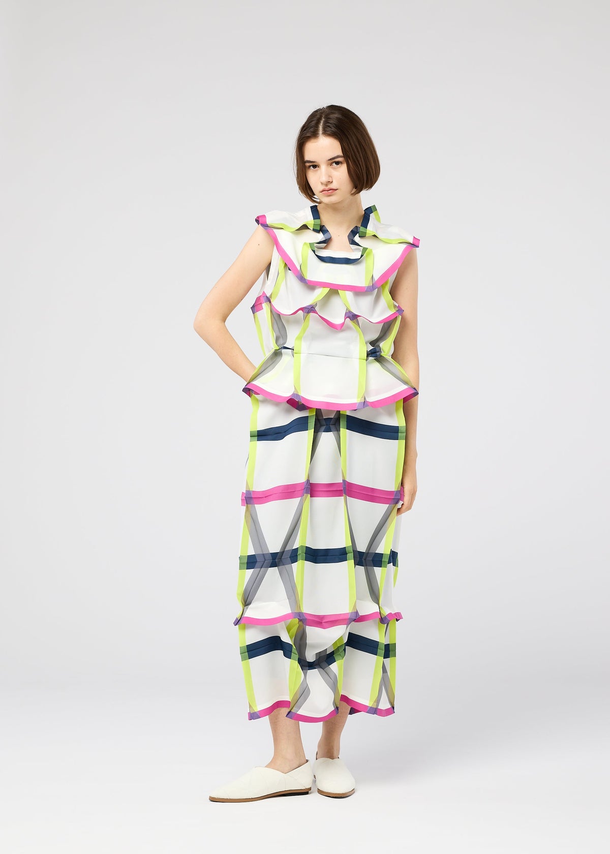 A woman wears the NO.6 TRANSLUCENT outfit by 132 5. ISSEY MIYAKE—loose, sleeveless top and wide-leg pants in a colorful grid print with sheer fabric—styled simply with white slippers against a plain backdrop and a neutral expression. view 11 of 11
