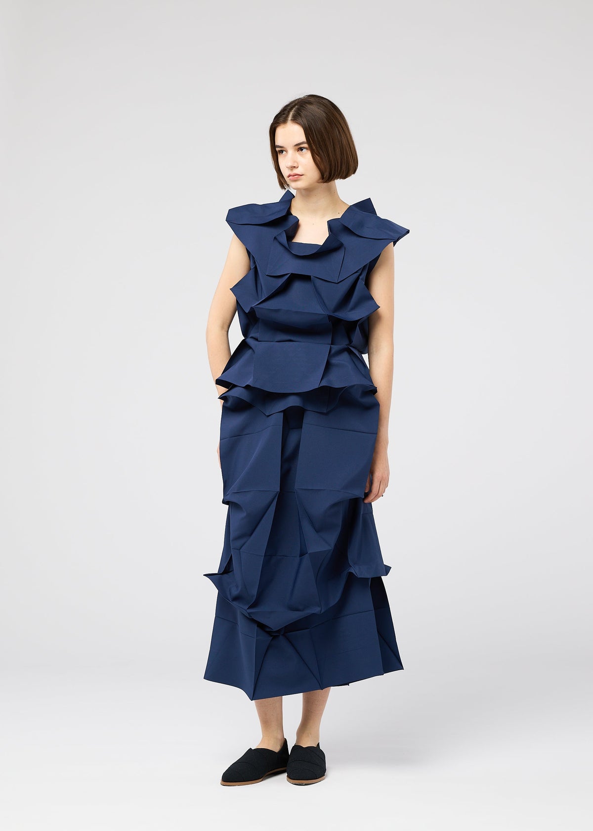 A woman with a straight bob wears the NO.6 SOLID dress by 132 5. ISSEY MIYAKE, featuring layered angular folds for a bold 3D look. She stands against a white background, gazing to the side with one hand in her pocket. view 5 of 9