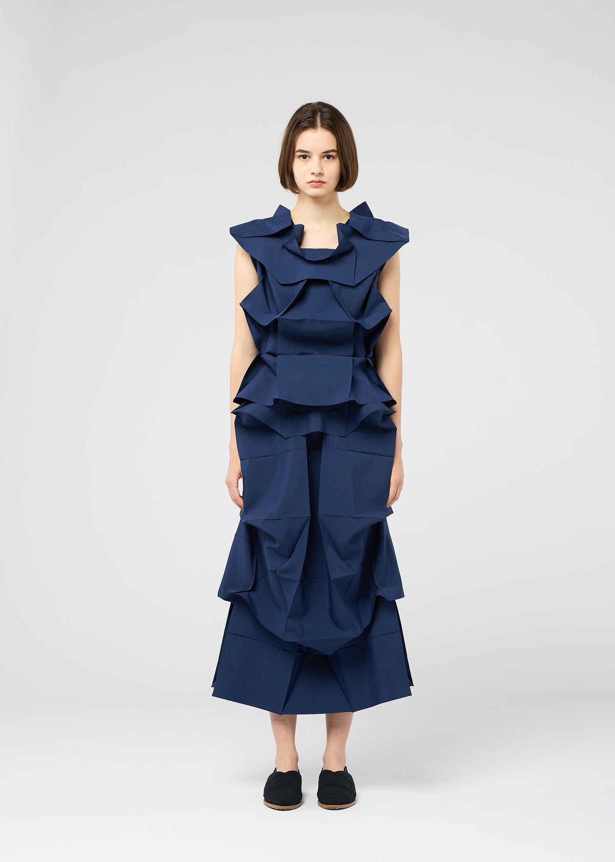 A woman with a neutral expression stands against a plain background, wearing the NO.6 SOLID dress from 132 5. ISSEY MIYAKE, featuring structured, navy fabric with geometric ruffles, paired with black slip-on shoes. view 6 of 9