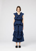 A woman with a neutral expression stands against a plain background, wearing the NO.6 SOLID dress from 132 5. ISSEY MIYAKE, featuring structured, navy fabric with geometric ruffles, paired with black slip-on shoes.