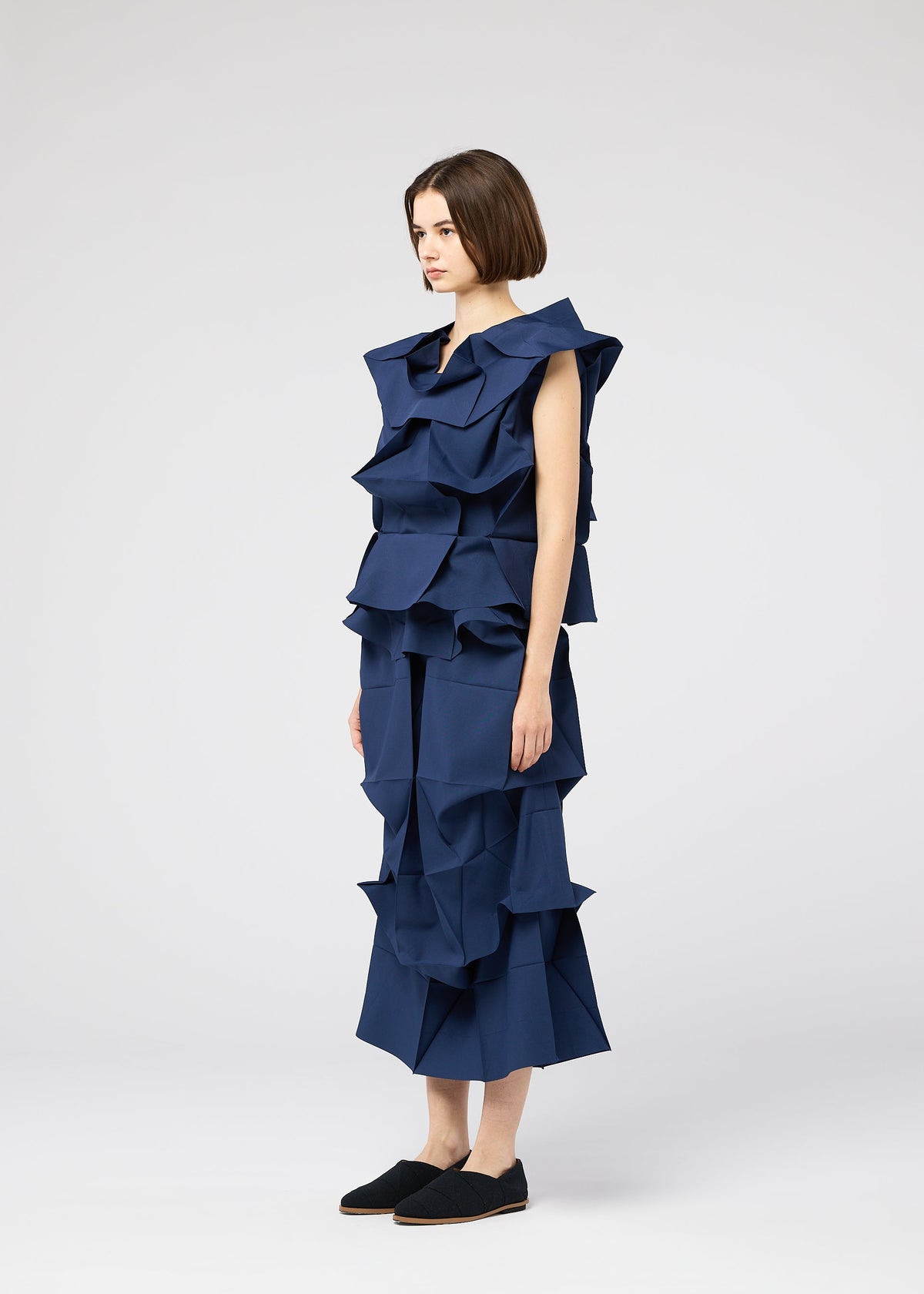 Against a plain background, a woman wears the NO.6 SOLID sleeveless dark blue dress by 132 5. ISSEY MIYAKE, with dramatic three-dimensional structure and origami-inspired ruffles, paired with black slip-on shoes and a short bob haircut. view 7 of 9