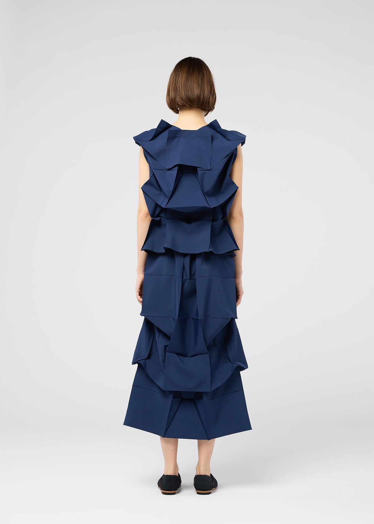 A person with short brown hair faces away, wearing the 132 5. ISSEY MIYAKE NO.6 SOLID sleeveless dark blue dress with layered, origami-style ruffles and black slip-on shoes, set against a plain white background. view 8 of 9