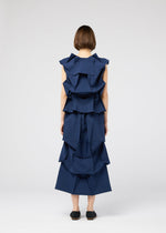 A person with short brown hair faces away, wearing the 132 5. ISSEY MIYAKE NO.6 SOLID sleeveless dark blue dress with layered, origami-style ruffles and black slip-on shoes, set against a plain white background.