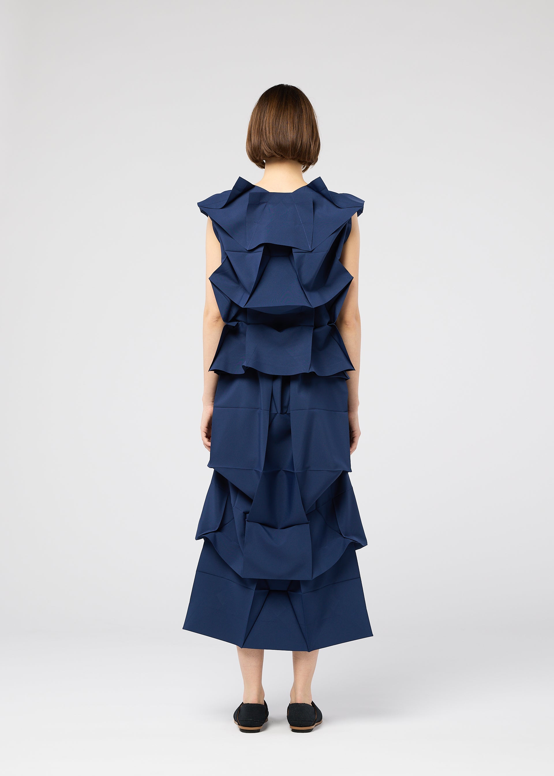 Zoomed view A person with short brown hair faces away, wearing the 132 5. ISSEY MIYAKE NO.6 SOLID sleeveless dark blue dress with layered, origami-style ruffles and black slip-on shoes, set against a plain white background.