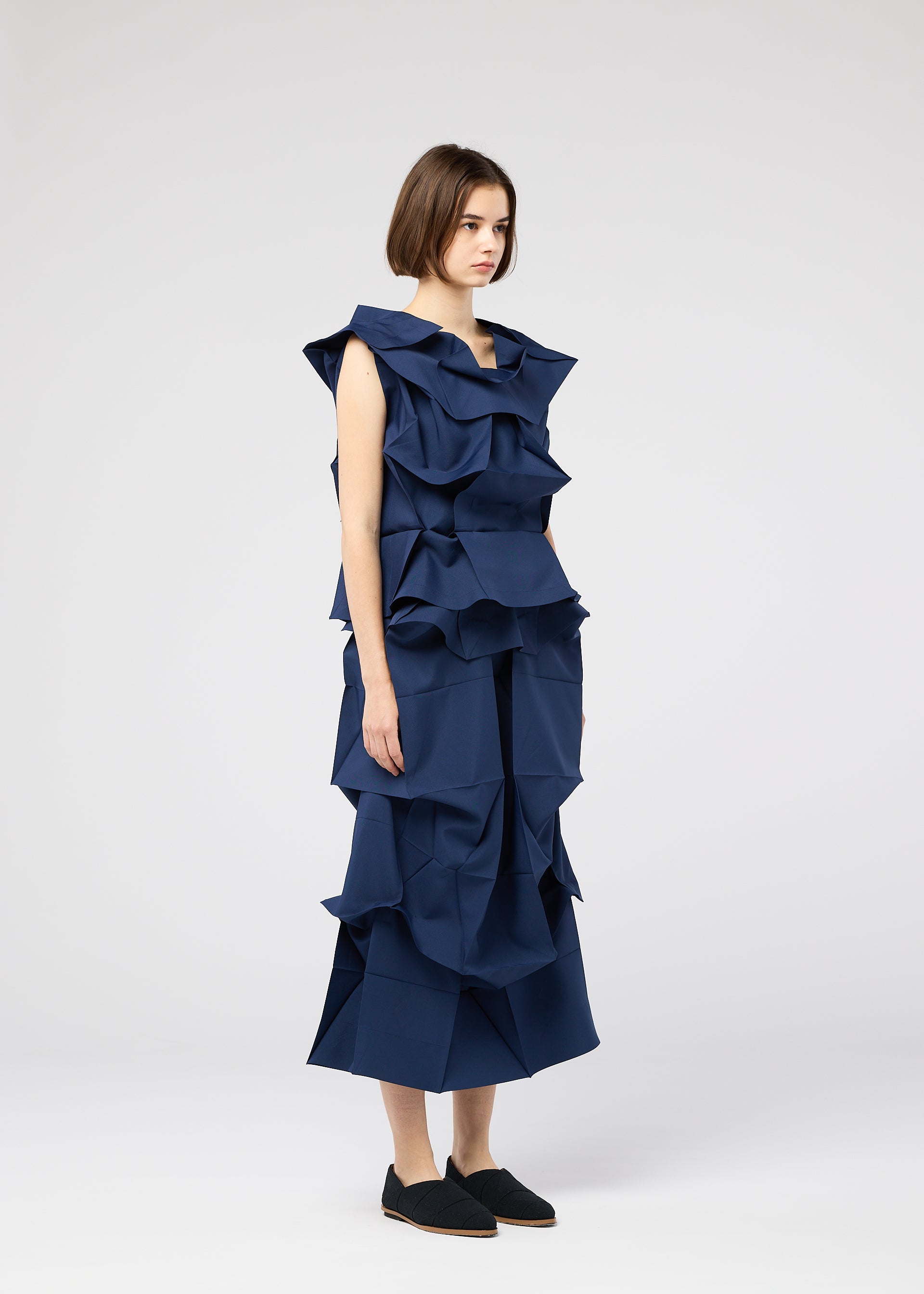 Zoomed view A woman stands against a white background wearing the NO.6 SOLID sleeveless navy dress with sculptural ruffles by 132 5. ISSEY MIYAKE, paired with black slip-on shoes.