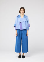 A woman with a straight brown bob faces forward, modeling the FOLD RAMIE PRINT jacket by 132 5. ISSEY MIYAKE in light blue and purple colors, paired with wide-legged blue pants and black slip-on shoes against a plain white backdrop.