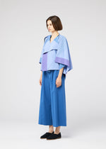 A woman with short brown hair stands against a plain background, wearing the FOLD RAMIE PRINT cape-style top in light blue and lavender by 132 5. ISSEY MIYAKE, wide-leg blue pants, and black shoes. She looks forward with a neutral expression.