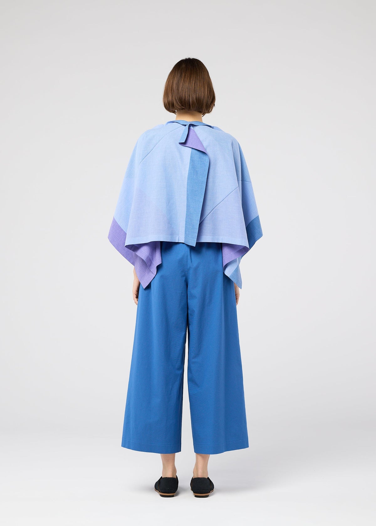 Against a plain white background, a person with short brown hair is seen from behind wearing the 132 5. ISSEY MIYAKE FOLD RAMIE PRINT: a light blue and purple cape-style top with unique color design and wide-legged blue pants. view 8 of 10