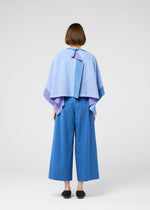 Against a plain white background, a person with short brown hair is seen from behind wearing the 132 5. ISSEY MIYAKE FOLD RAMIE PRINT: a light blue and purple cape-style top with unique color design and wide-legged blue pants.