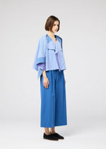 A woman with short brown hair wears the FOLD RAMIE PRINT by 132 5. ISSEY MIYAKE—a light blue layered jacket over a blue top and wide-leg pants—standing sideways against a plain light gray background, finished with black slip-on shoes.