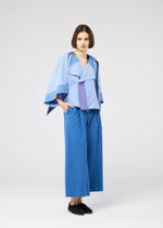A woman with short brown hair wears the FOLD RAMIE PRINT jacket by 132 5. ISSEY MIYAKE, featuring a loose geometric blue and lavender design over wide-legged blue pants and black slip-ons, standing hands-in-pockets against a white background.