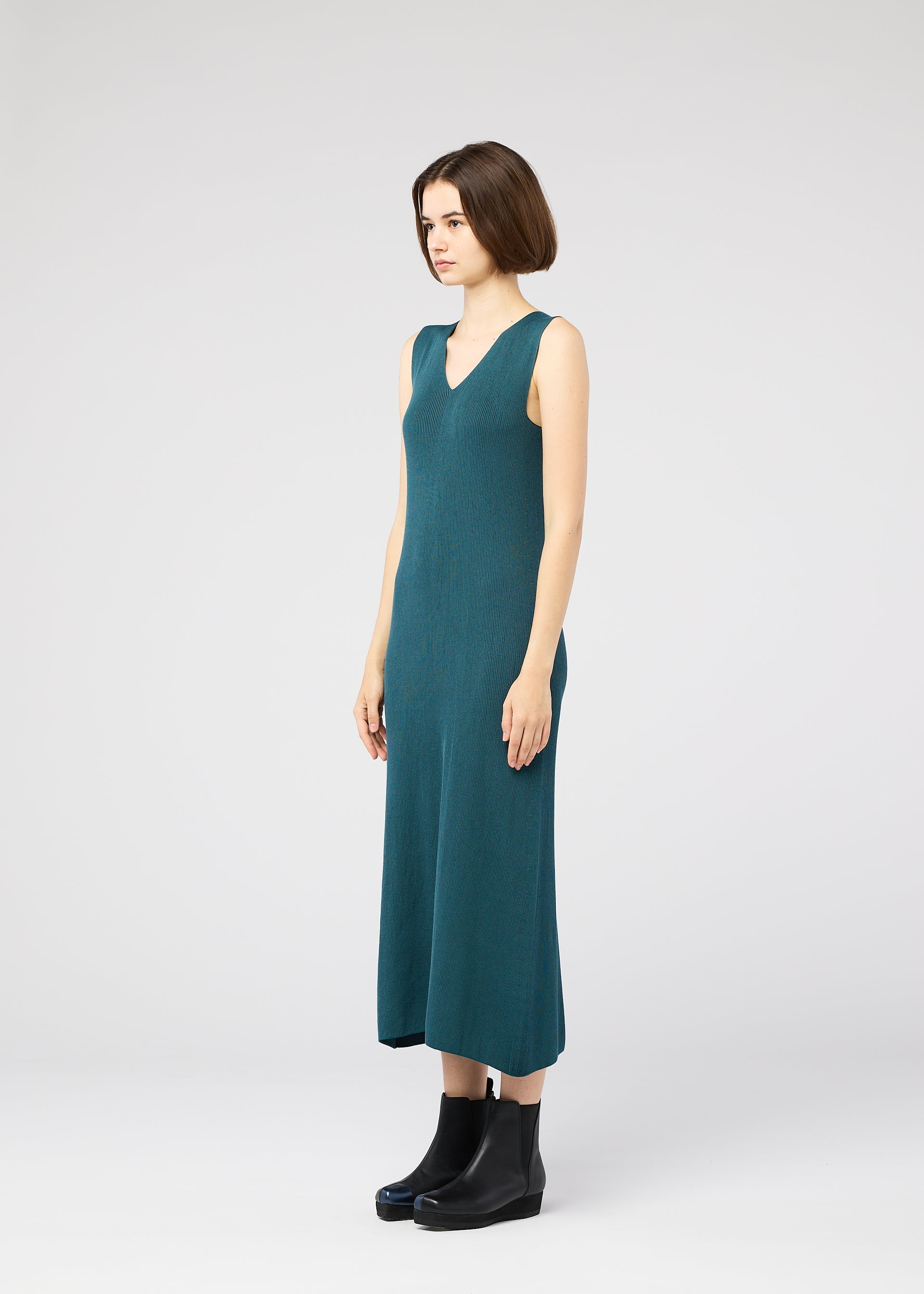 Zoomed view A woman with a bob haircut models the 132 5. ISSEY MIYAKE FLAT RIB KNIT 1, a sleeveless teal dress, paired with black boots. She stands against a plain light gray background, facing slightly left.