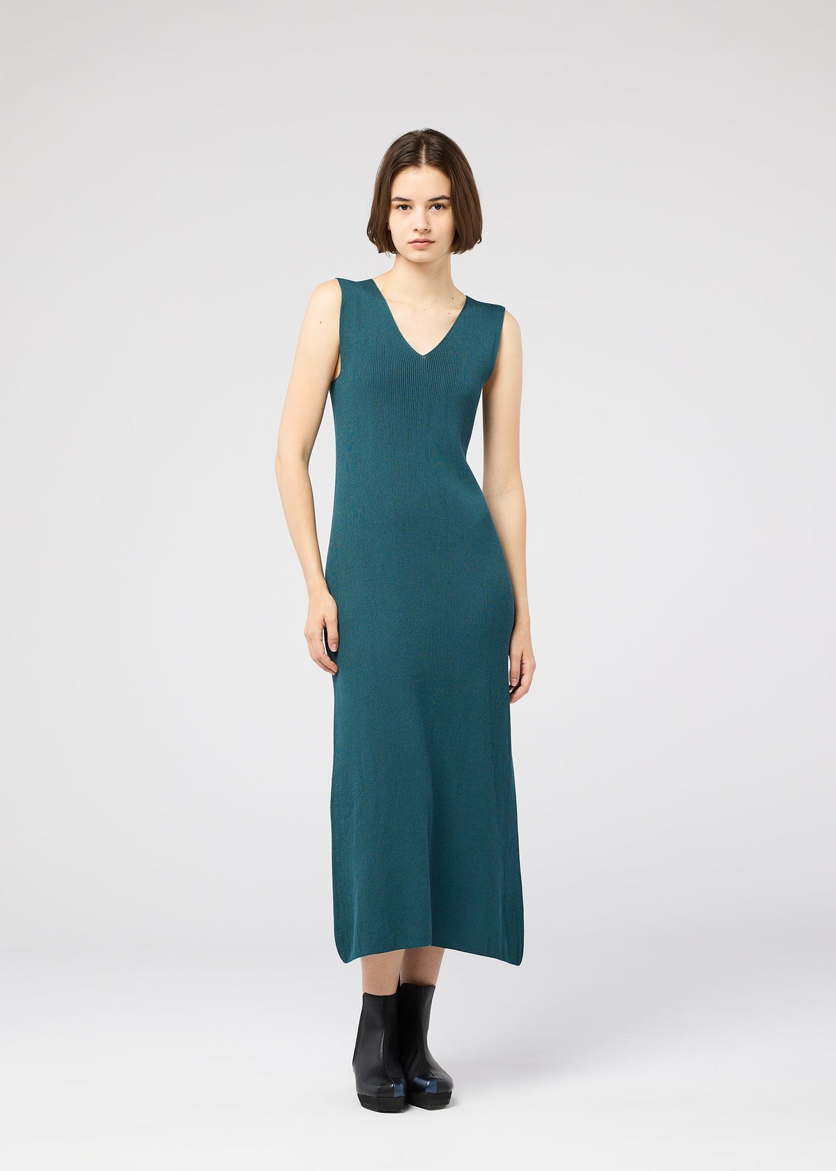 A woman with a short brown bob models the 132 5. ISSEY MIYAKE FLAT RIB KNIT 1, a sleeveless teal V-neck seamless rib knit dress, paired with black ankle boots, standing against a plain light gray background and facing forward. view 10 of 10