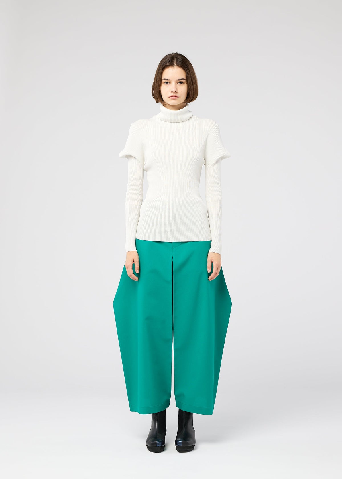 A woman with a bob haircut wears the FLAT RIB KNIT 1 seamless white turtleneck sweater by 132 5. ISSEY MIYAKE and wide teal-green pants, standing against a plain light gray background, facing forward. view 7 of 10