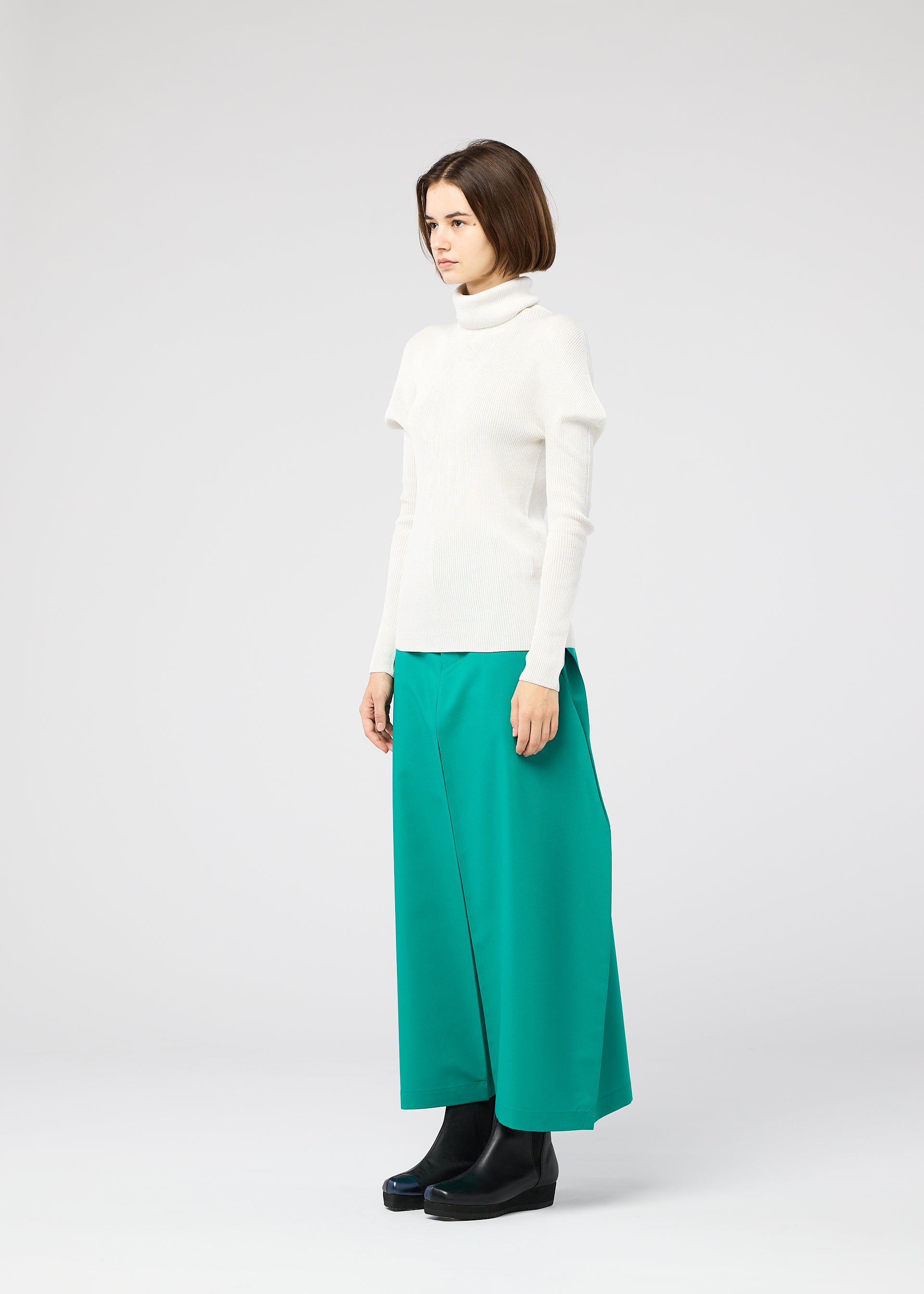 Zoomed view A woman in profile wears the 132 5. ISSEY MIYAKE FLAT RIB KNIT 1 white turtleneck sweater, wide-legged green trousers, and black platform shoes against a plain light gray background.