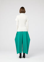 A person with short brown hair faces away, wearing the 132 5. ISSEY MIYAKE FLAT RIB KNIT 1 white turtleneck and wide, bright green geometric pants with black ankle boots, set against a simple light background.