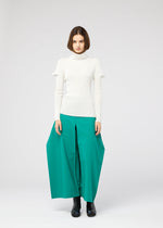 A woman stands facing forward, wearing the 132 5. ISSEY MIYAKE FLAT RIB KNIT 1 white turtleneck sweater, a wide bright teal skirt with front slits, and black boots, against a plain light gray background.