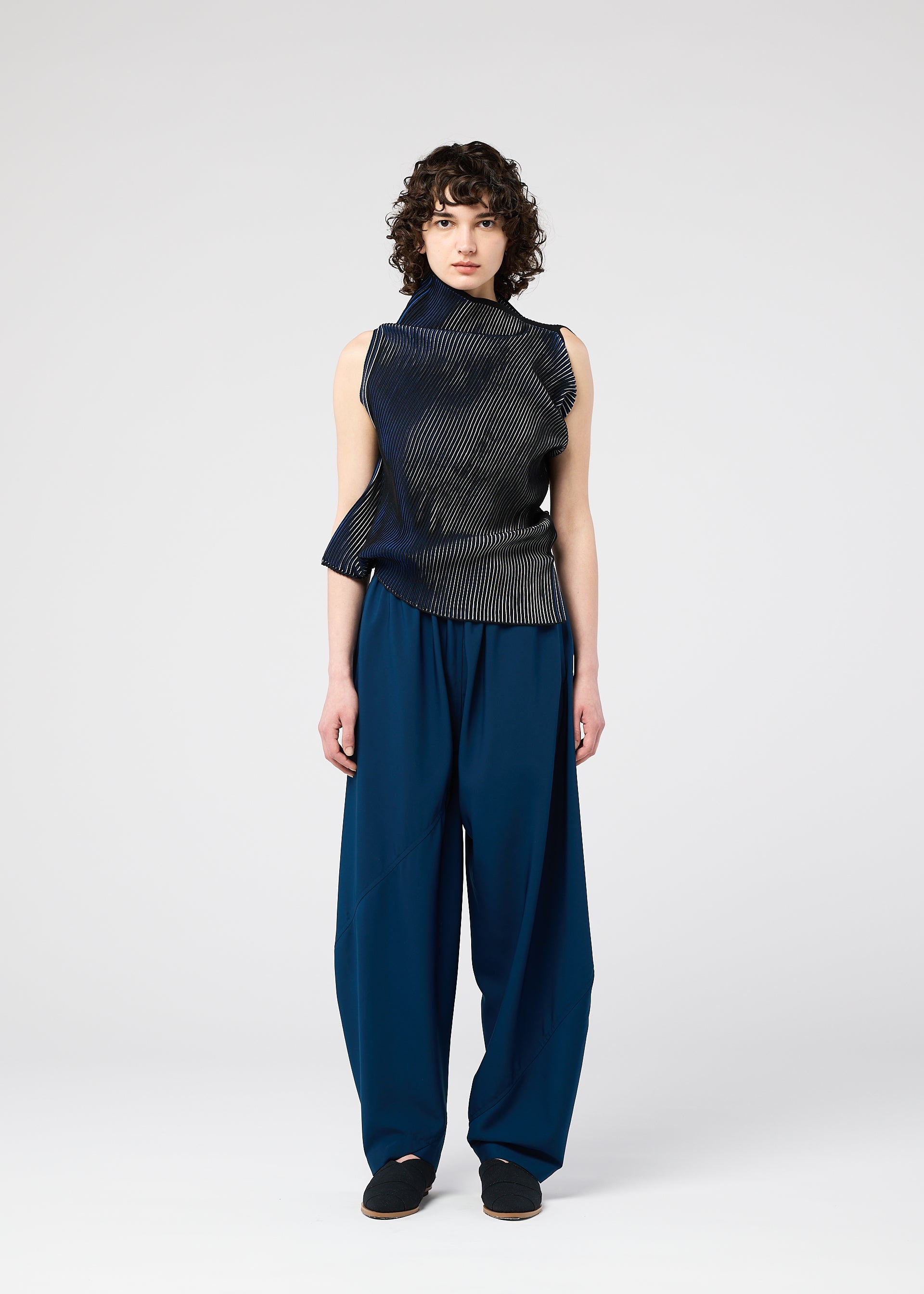 Zoomed view A person with short curly hair stands facing forward, wearing the FACET KNIT sleeveless asymmetrical dark top by 132 5. ISSEY MIYAKE, featuring a striped pattern and loose blue pants. The background is plain light gray.