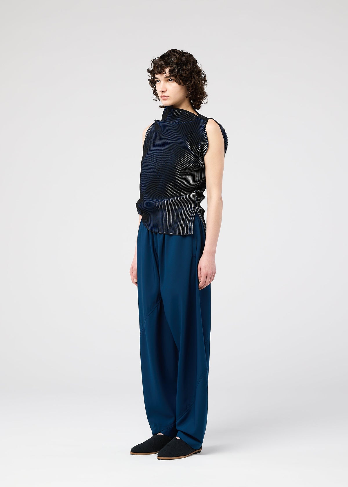 A person with curly hair wears the FACET KNIT sleeveless top by 132 5. ISSEY MIYAKE, featuring a unique textured knit, paired with loose blue trousers and standing against a plain light background, facing slightly right. view 7 of 11