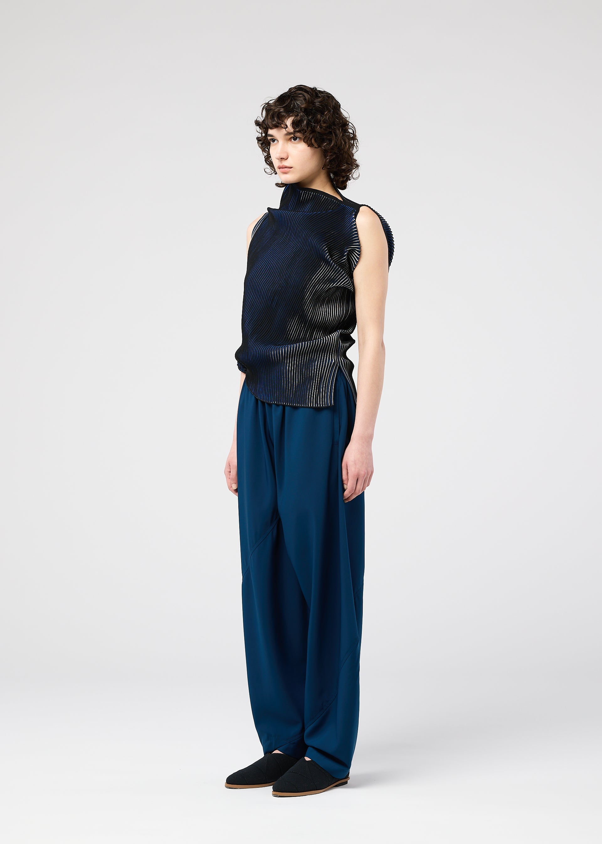 Zoomed view A person with curly hair wears the FACET KNIT sleeveless top by 132 5. ISSEY MIYAKE, featuring a unique textured knit, paired with loose blue trousers and standing against a plain light background, facing slightly right.