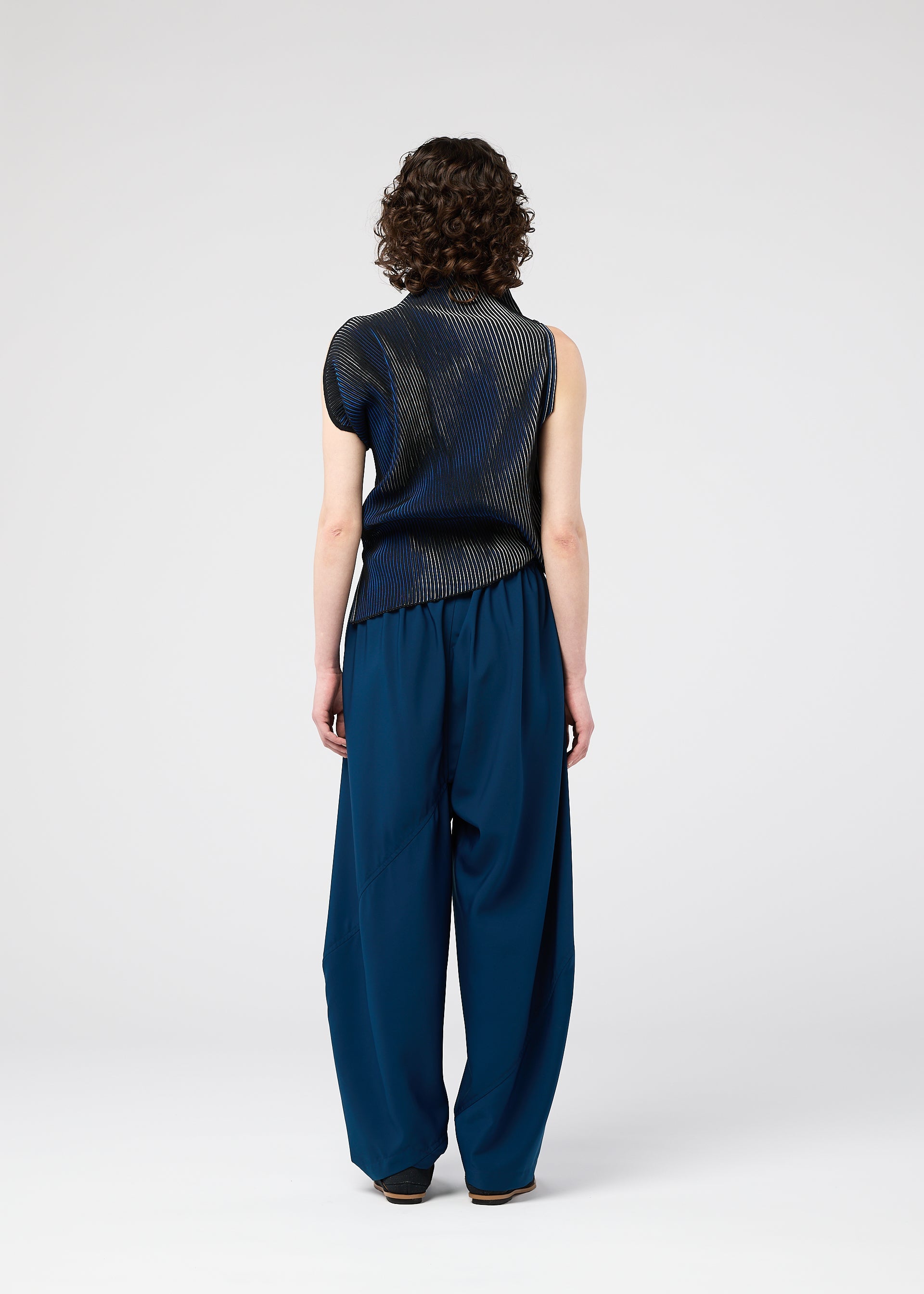 Zoomed view Seen from behind, a person with short curly hair wears the 132 5. ISSEY MIYAKE FACET KNIT sleeveless dark navy top and loose, wide-legged blue pants, standing against a plain light background.