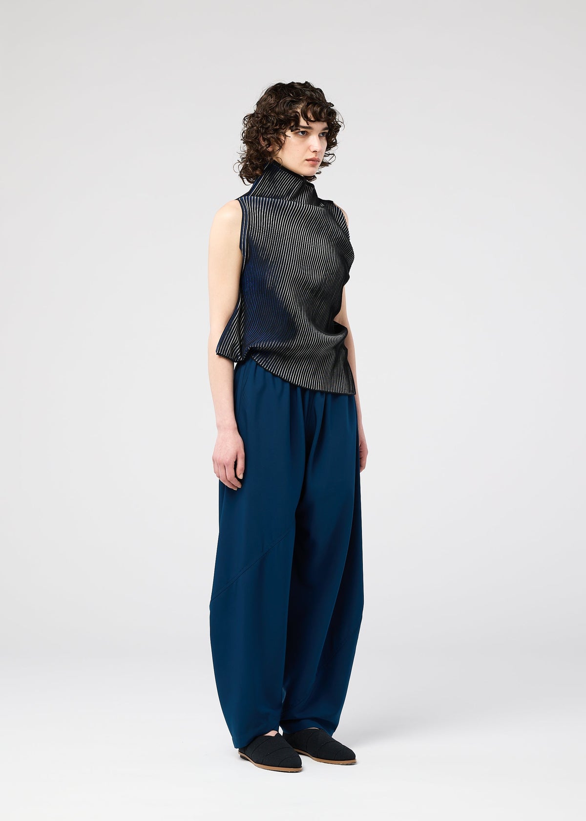 A person with short curly hair wears the FACET KNIT sleeveless black top by 132 5. ISSEY MIYAKE and loose-fitting, high-waisted blue pants, standing against a plain light background. view 9 of 11