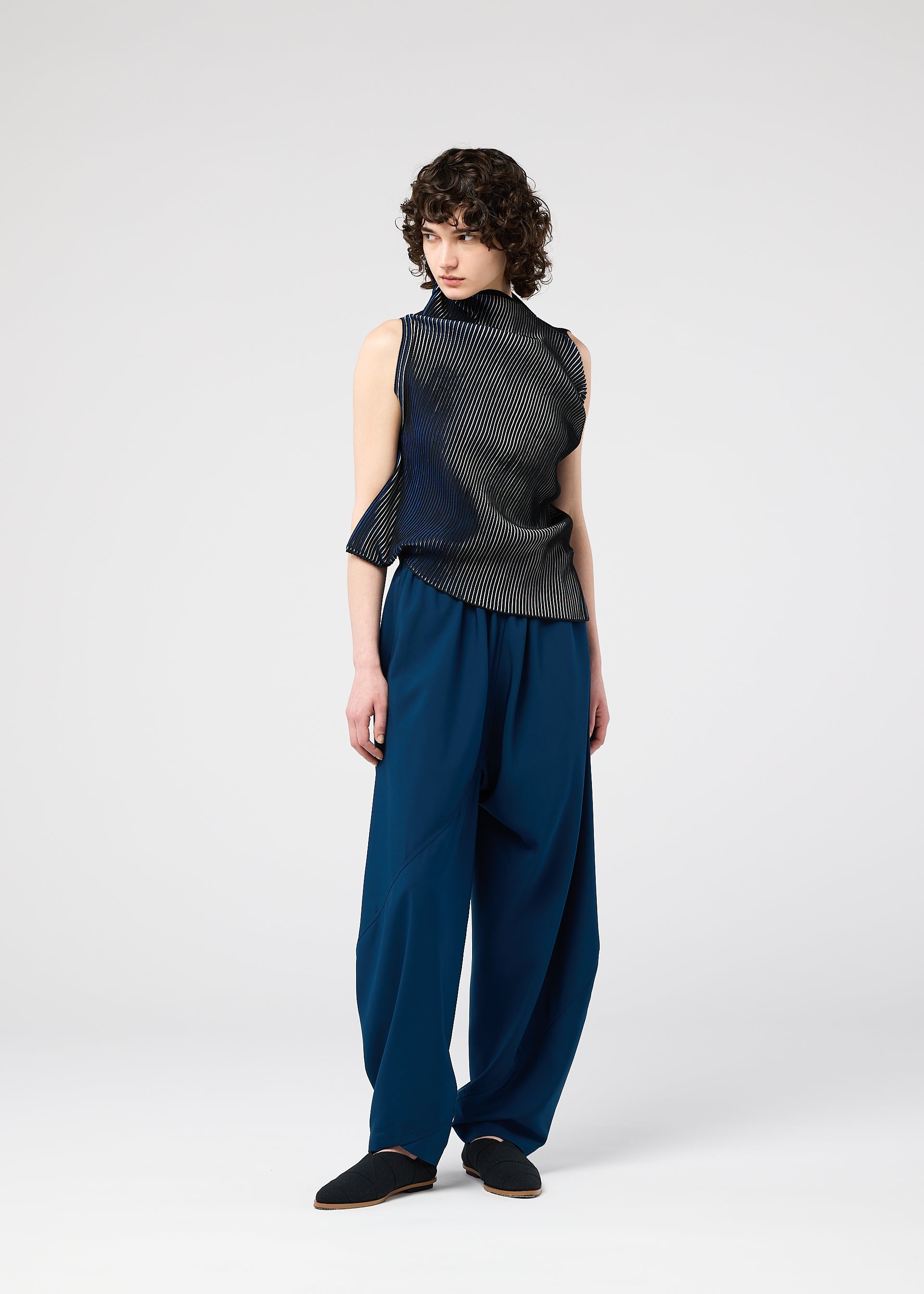 Zoomed view A person with short curly hair wears the FACET KNIT by 132 5. ISSEY MIYAKE—a sleeveless, asymmetrical black-and-white striped top—paired with loose blue pants, standing against a plain light background.