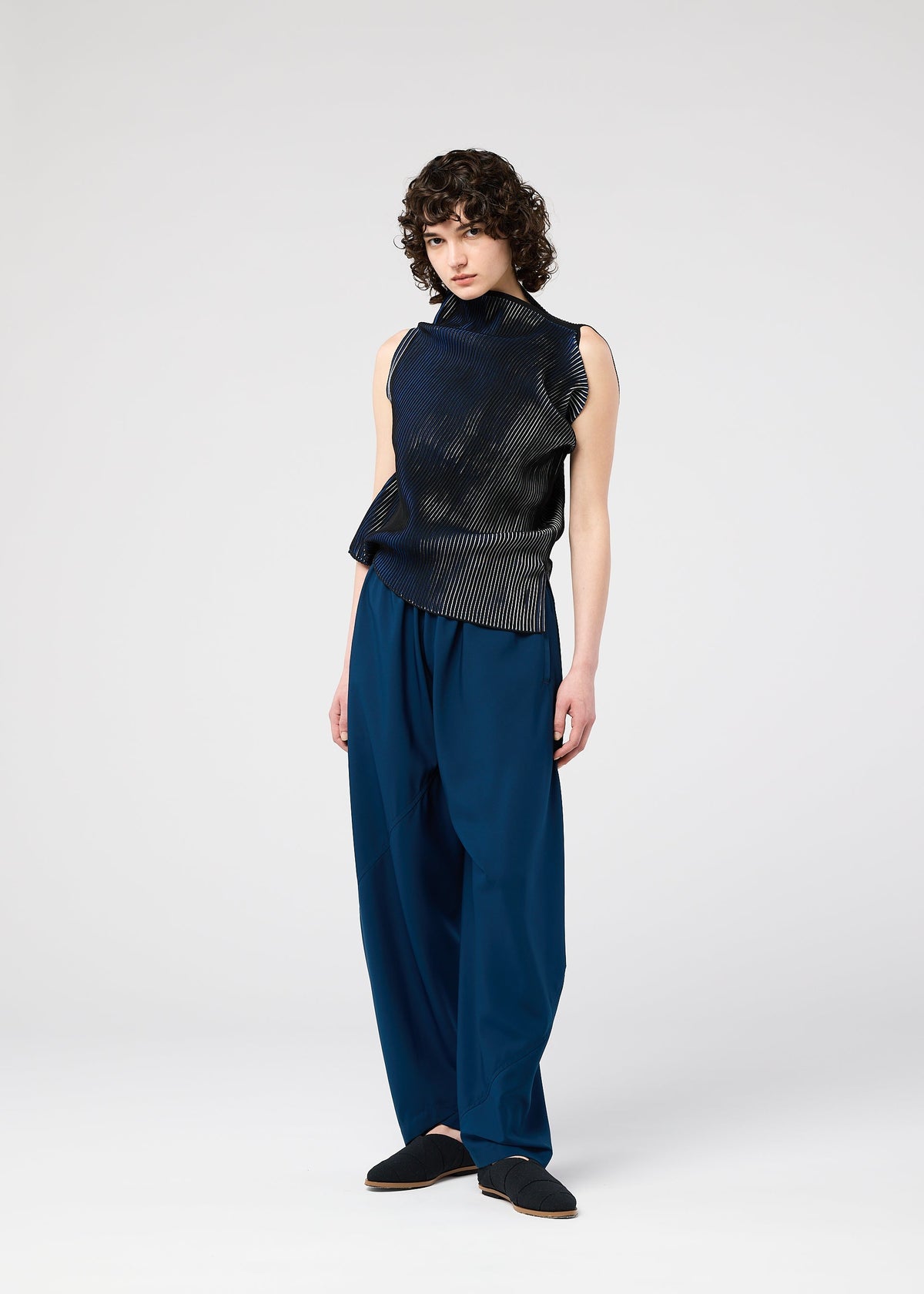 A person with short curly hair wears the FACET KNIT by 132 5. ISSEY MIYAKE—a sleeveless, asymmetrical black top with a special knit structure and subtle sheen—paired with loose blue pants, against a plain light background. view 11 of 11