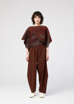 A person with short, curly hair and a neutral expression stands against a plain white background, wearing the 132 5. ISSEY MIYAKE FACET KNIT in maroon with brown wide-leg pants and white shoes. The top features unique textured patterns.