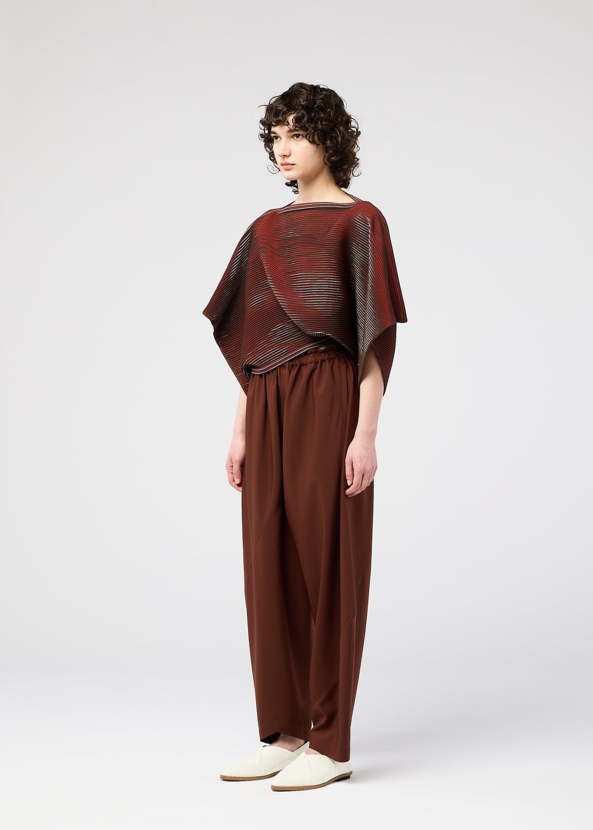 A person with curly hair stands sideways against a plain white background, wearing wide-legged brown pants, light-colored shoes, and a loose red and brown striped FACET KNIT top from 132 5. ISSEY MIYAKE, known for its unique knit structure and pattern depth. view 6 of 10