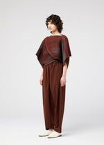 A person with curly hair stands sideways against a plain white background, wearing wide-legged brown pants, light-colored shoes, and a loose red and brown striped FACET KNIT top from 132 5. ISSEY MIYAKE, known for its unique knit structure and pattern depth.
