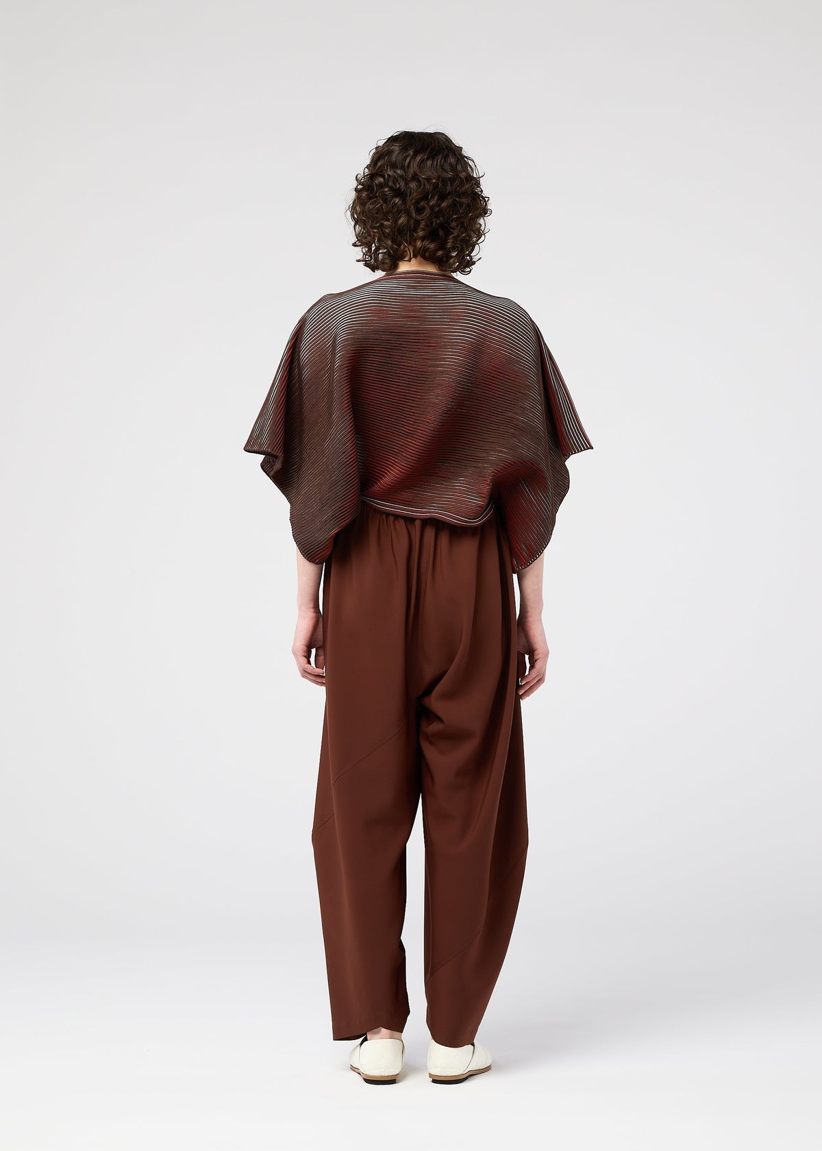 A person with curly hair stands facing away, wearing the 132 5. ISSEY MIYAKE FACET KNIT—a loose, striped brown-gray top—paired with high-waisted brown trousers and white shoes, set against a plain light background. view 7 of 10