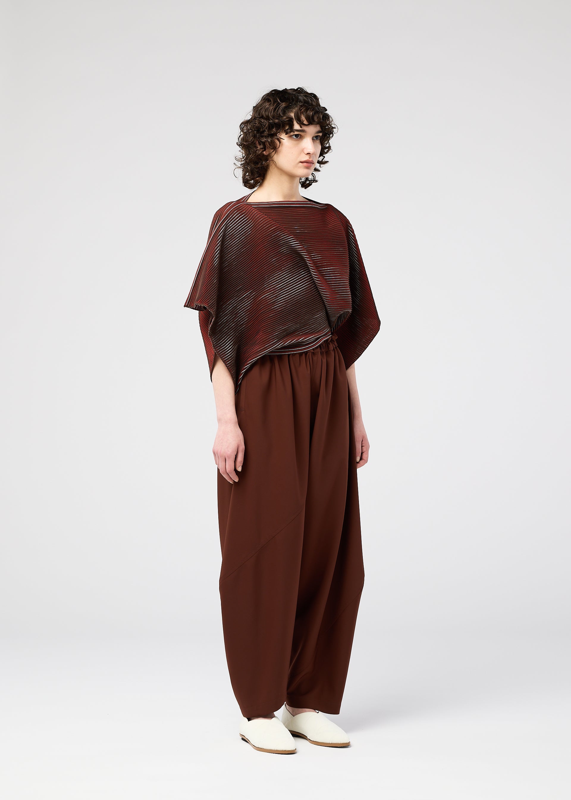 Zoomed view A person with curly hair stands against a plain background, wearing the FACET KNIT top and matching wide pants by 132 5. ISSEY MIYAKE, highlighting the unique knitting structure and pattern depth.