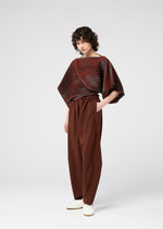 A person with short curly hair wears the FACET KNIT by 132 5. ISSEY MIYAKE, a loose, shiny maroon top with pattern depth and wide sleeves, paired with high-waisted brown pants and white shoes against a plain light gray background.