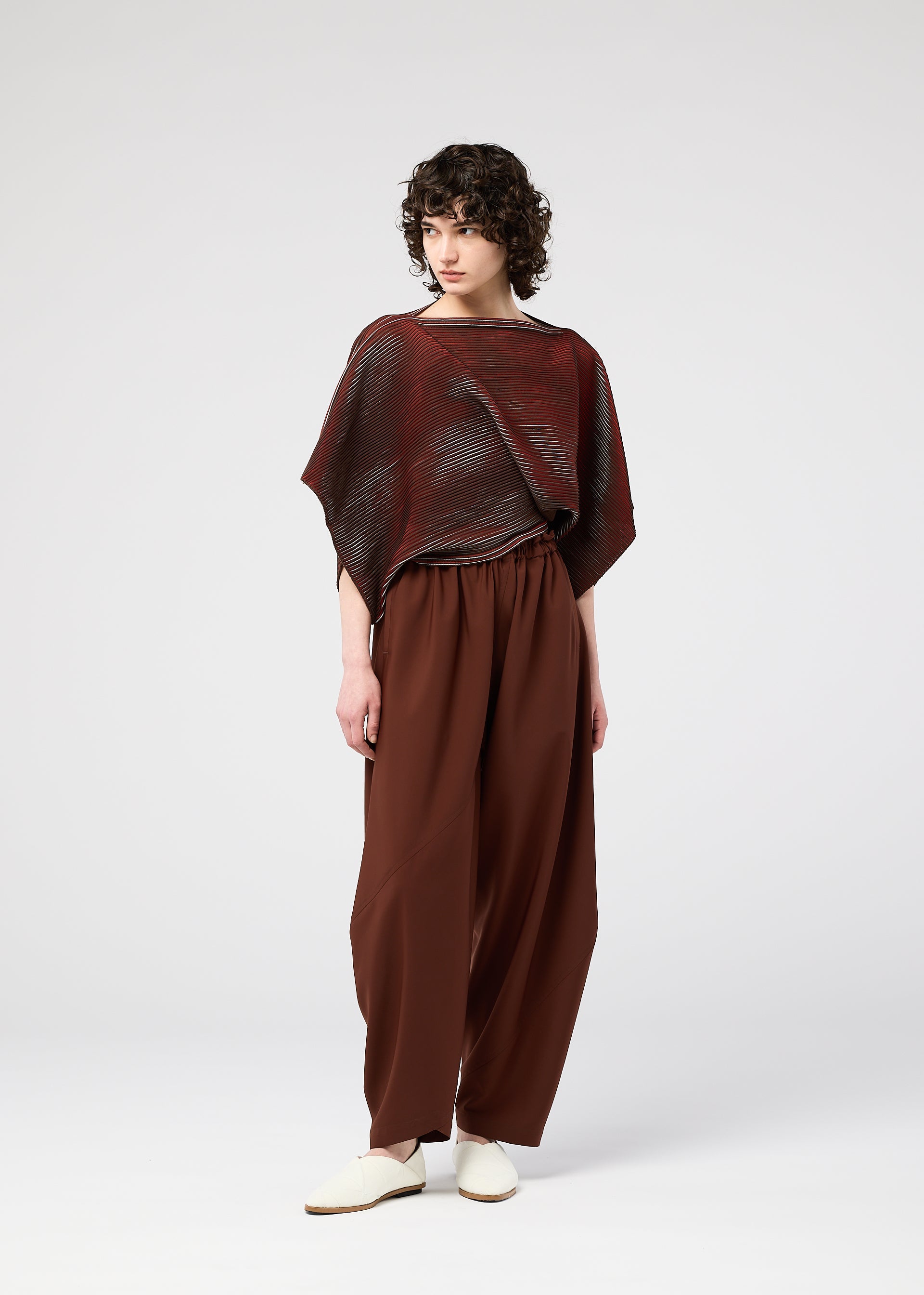 Zoomed view A person with short curly hair wears the FACET KNIT top by 132 5. ISSEY MIYAKE—a loose, semi-sheer maroon piece with textured pattern—paired with brown wide-leg pants and white slip-on shoes, standing against a plain light gray background.