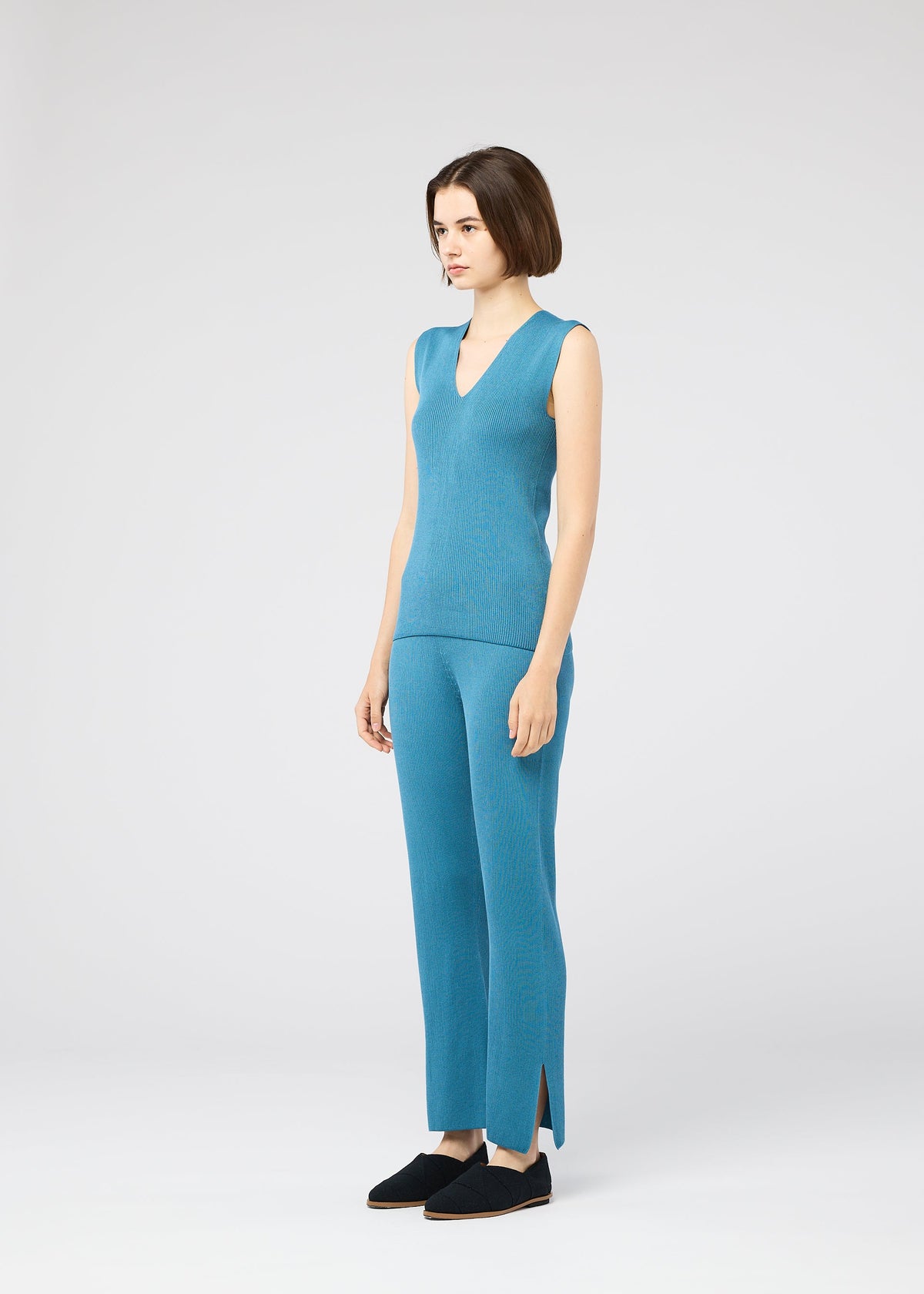 A woman with a short bob stands in a studio wearing the FLAT RIB KNIT 2 top and matching pants by 132 5. ISSEY MIYAKE—sleeveless, seamless blue rib knit with side-slit hems—paired with black slip-on shoes, facing slightly to the side. view 7 of 9
