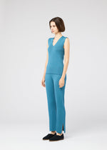 A woman with a short bob stands in a studio wearing the FLAT RIB KNIT 2 top and matching pants by 132 5. ISSEY MIYAKE—sleeveless, seamless blue rib knit with side-slit hems—paired with black slip-on shoes, facing slightly to the side.