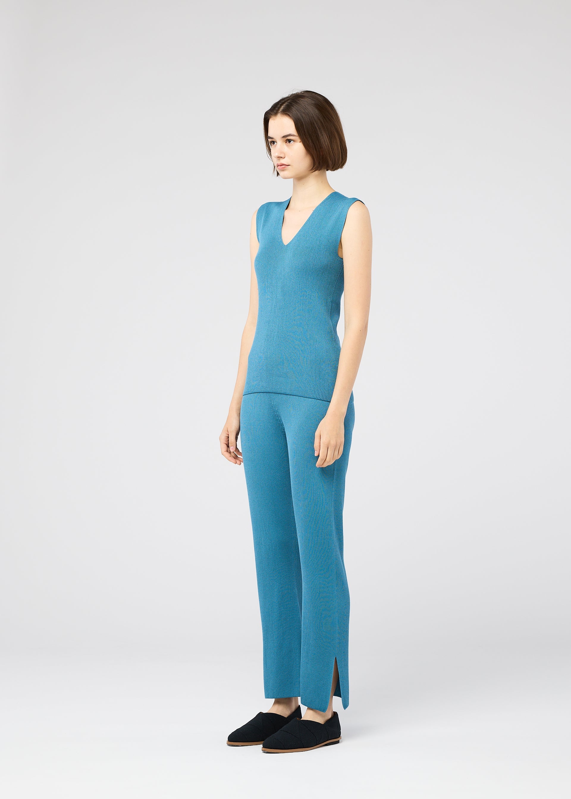Zoomed view A woman with a short bob stands in a studio wearing the FLAT RIB KNIT 2 top and matching pants by 132 5. ISSEY MIYAKE—sleeveless, seamless blue rib knit with side-slit hems—paired with black slip-on shoes, facing slightly to the side.